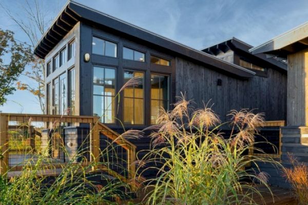 A modern wooden house with large windows, elevated deck, and surrounded by grassy plants under a clear sky. The structure has a slanted roof.