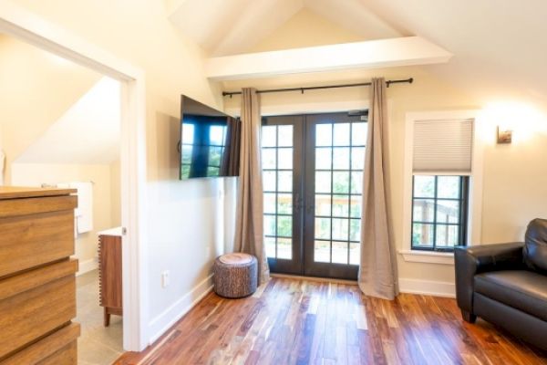 A small, modern room with a flat-screen TV, armchair, chest of drawers, French doors, wooden floors, and beige walls.