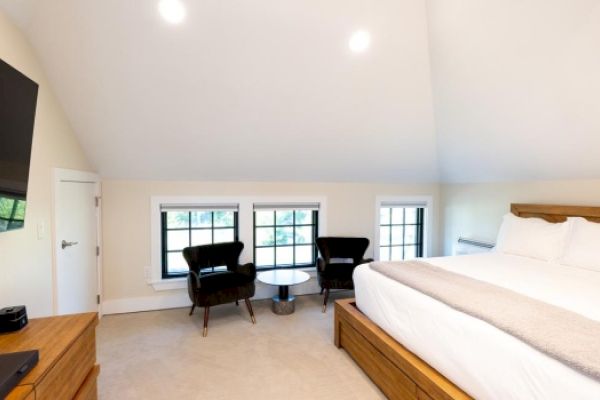 A cozy bedroom with a large bed, two chairs by the windows, a small table, a wooden dresser, and a TV mounted on the wall.