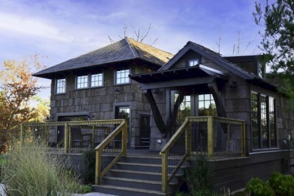 A two-story house featuring rustic wooden finishes, nestled in a natural setting, and surrounded by greenery under a blue sky, is shown.