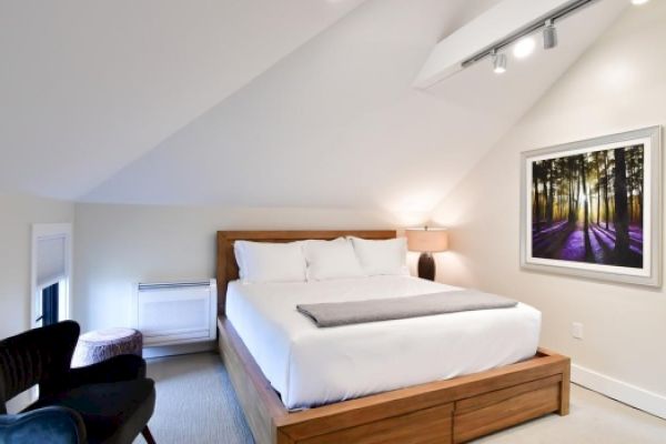 A cozy bedroom with sloped ceilings, a bed with white linens, a nightstand with a lamp, a chair, a small table, and a framed forest picture.