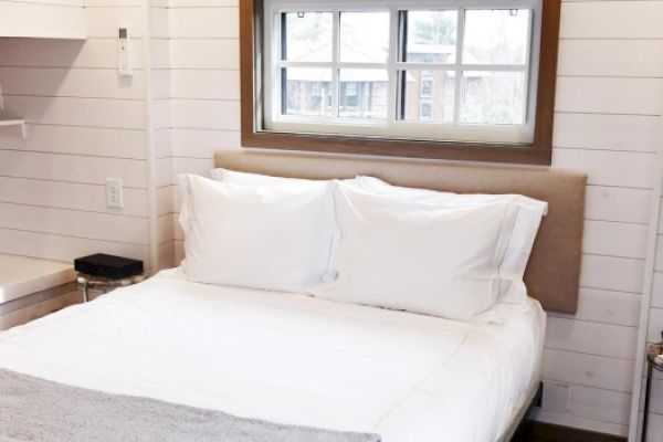 A cozy bedroom with white walls, a double bed with white bedding, a small window above the bed, and minimalistic furniture on either side.