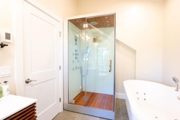 A modern bathroom with a glass shower enclosure, a white bathtub, a mirror, and a white door on the left.