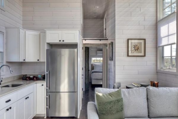 The image shows a modern, small living space with white wooden walls, a kitchen to the left, a stainless steel fridge, and a gray couch on the right, ending the sentence.