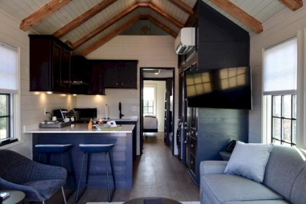 A modern tiny house interior with a lofted ceiling, featuring a kitchen, bar stools, living area with sofa, TV, and a bedroom in the back.