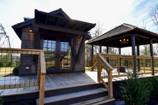 The image shows a small wooden cabin with a porch and a separate gazebo beside it. The structures are surrounded by trees and shrubs.