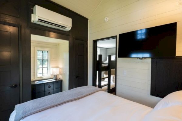 A cozy bedroom features a large bed, wall-mounted TV, air conditioner, dark wooden accents, and an adjoining room with bunk beds.