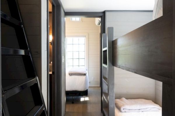 The image shows an interior hallway with a dark wooden bunk bed on one side and a door leading to a bright room with a bed and window.