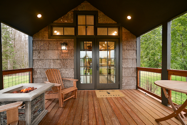 A cozy porch with wooden flooring, a table, chairs, a fire pit, and glass door entry, surrounded by trees and greenery in the background.