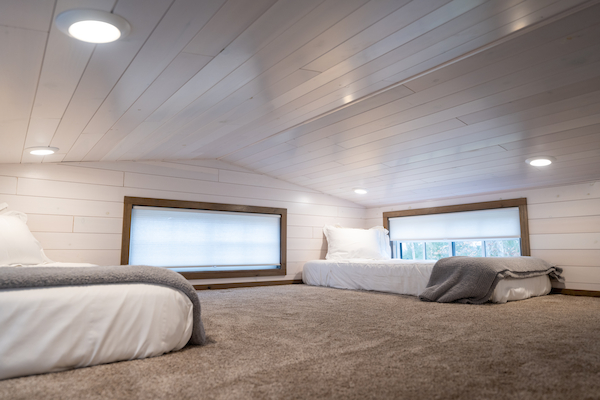 This image shows a cozy loft space with a low ceiling, featuring two beds with white bedding and blankets, carpeted floor, and small windows.