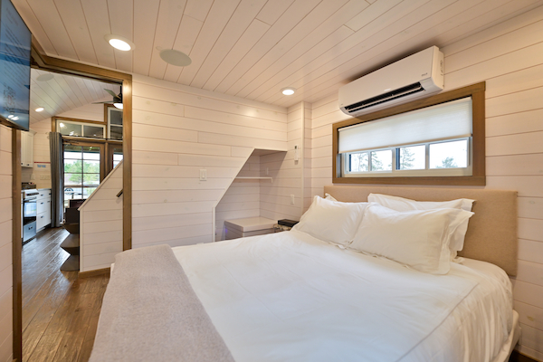 A cozy bedroom with white walls, a double bed, air conditioner, and a small desk space. There's a staircase leading to another area.