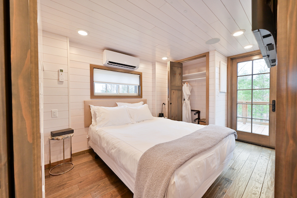 A cozy bedroom with white walls, wood floor, bed with white linens, wall-mounted TV, air conditioner, side tables, and a door to the balcony.