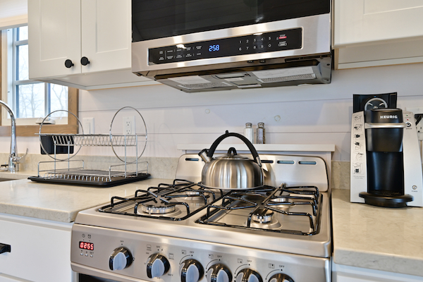 A kitchen with a gas stove, teapot, dish rack, microwave, coffee maker, and white cabinets. A window can be seen on the left.