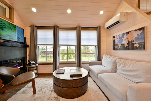 A cozy living room with a light grey sofa, round wooden coffee table, wall-mounted TV, large windows with curtains, and contemporary decor.