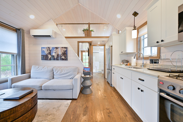 A cozy tiny house interior with a couch, artwork, kitchenette, and bathroom. The design features white walls, wood flooring, and ample lighting.