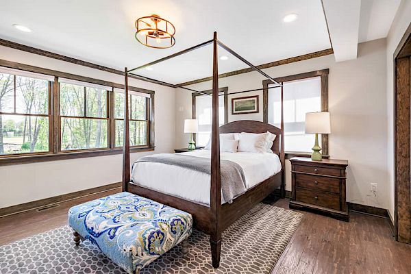 A cozy bedroom with a canopy bed, patterned bench, large windows, and wooden furniture, including nightstands with lamps, is shown.