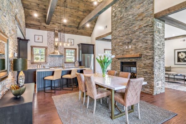 A modern open-concept living space with a kitchen, dining area, and living room, featuring wooden beams, stone walls, and stylish furnishings.