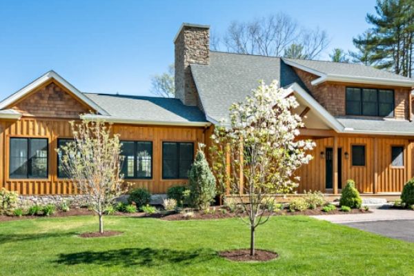 A modern two-story house with wood siding, large windows, and a stone chimney, surrounded by a well-manicured lawn, trees, and landscaping.