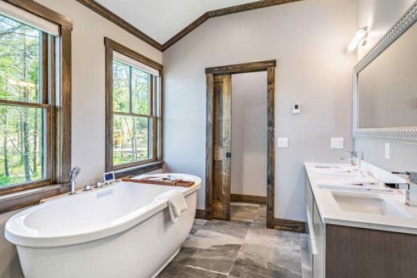 This image shows a modern bathroom with a freestanding bathtub, double sink vanity, large mirror, and two windows with wooded views outside.