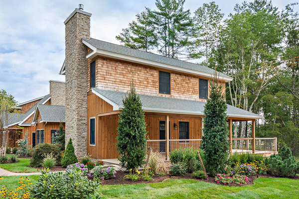 A rustic two-story house with wooden siding, a tall stone chimney, surrounded by greenery and colorful flowers, set against a backdrop of trees.