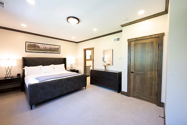 A modern bedroom with a large bed, two nightstands with lamps, a dresser with a mirror, and wooden doors on light-colored walls and carpet.