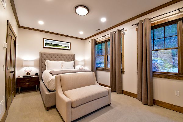 This image shows a cozy bedroom with a bed, nightstands with lamps, a bench at the foot of the bed, and two windows with beige curtains.