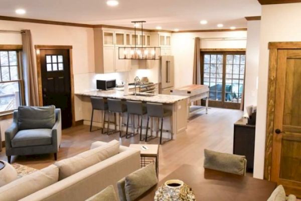 A modern open-plan kitchen and living room featuring a stone fireplace, island with barstools, and a mix of wooden and white cabinets.