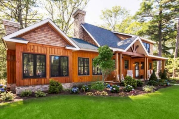 A charming wooden house surrounded by lush greenery, featuring large windows, a porch with rocking chairs, and a well-manicured lawn.