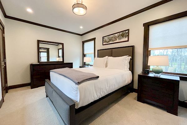 A neatly arranged modern bedroom with a bed, bedside tables, lamps, and a dresser with a mirror. The room features neutral colors.