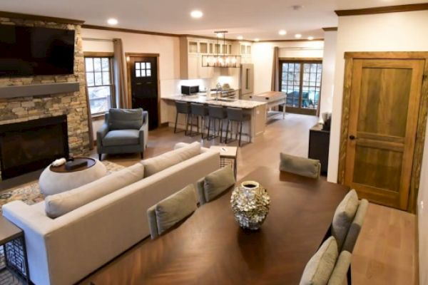 A modern living room with a stone fireplace, kitchen with island seating, dining table with chairs, and wooden floor.