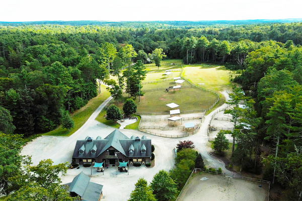 Aerial view of a large facility surrounded by forest, featuring multiple buildings, fenced areas, pathways, and a main building at the center of the facility.