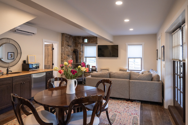 This image shows a cozy living room and kitchen area with a dining table, sofas, a TV, and flowers on the table. The room appears well-lit.