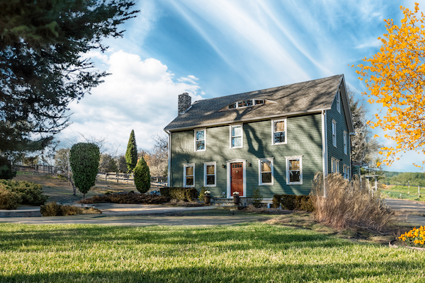 The image shows a two-story green house with a brown roof, surrounded by a well-maintained lawn, trees, and vibrant autumn foliage under a blue sky.