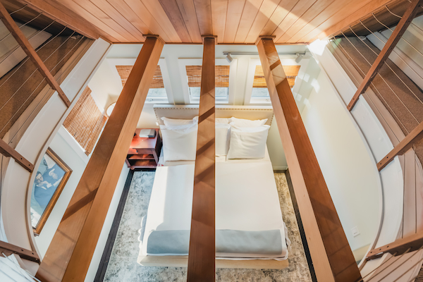 The image shows an aerial view of a bedroom with a white bed, wooden beams crossing above, a bedside table, a framed picture, and windows.