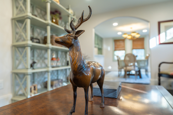 A bronze deer sculpture stands on a wooden table with a living room and dining area in the background, under warm lighting.