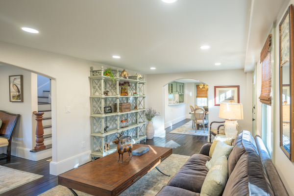 This image shows a cozy living room with a sofa, coffee table, shelves, and various decor, connected to a dining area, well-lit by ceiling lights.