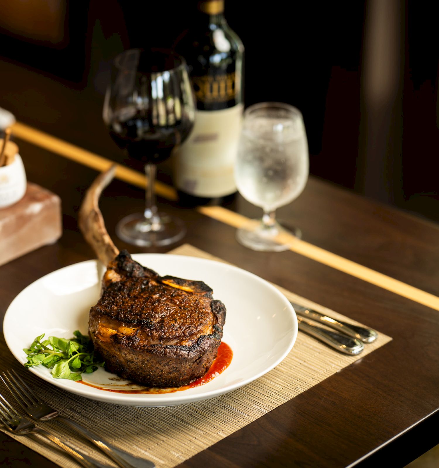 The image shows a cooked tomahawk steak on a white plate with garnish, alongside a glass of red wine, a glass of water, and seasoning containers.