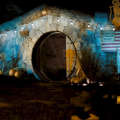 A circular stone entrance at night is illuminated with string lights. An American flag and a sign that reads 