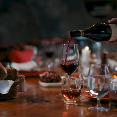 A person pouring wine into a glass on a dinner table set with food, candles, and various drinks in glasses.