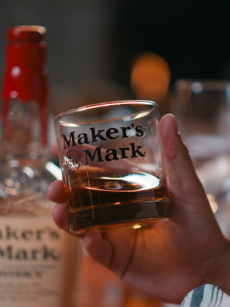 A hand holds a glass of Maker's Mark whiskey, with several Maker's Mark bottles visible in the background on a wooden surface.