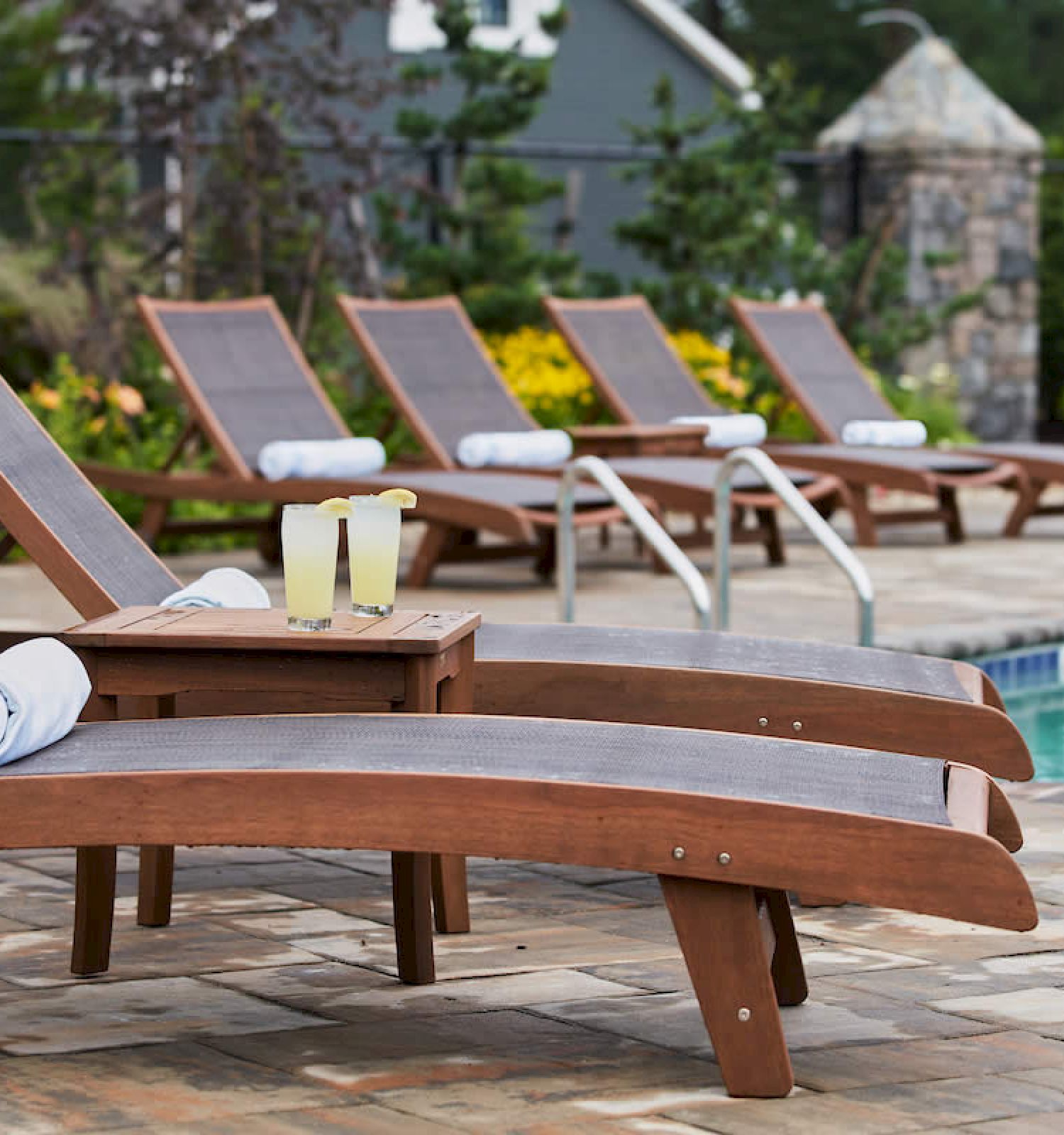 Poolside lounge chairs with rolled-up towels and a small table holding two drinks, set beside a swimming pool in an outdoor setting.