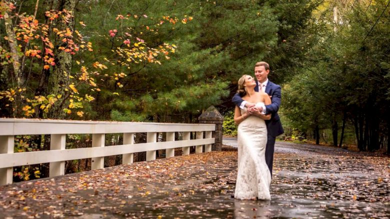 A couple dressed in wedding attire embraces and smiles on a wet road surrounded by autumn foliage, next to a white fence.