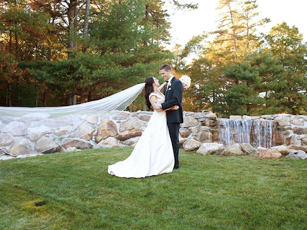 A couple in wedding attire embrace outdoors near a stone waterfall, surrounded by trees and grass.