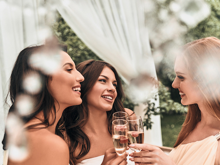 Three women are smiling and clinking champagne glasses in a festive outdoor setting, surrounded by white drapes and greenery.