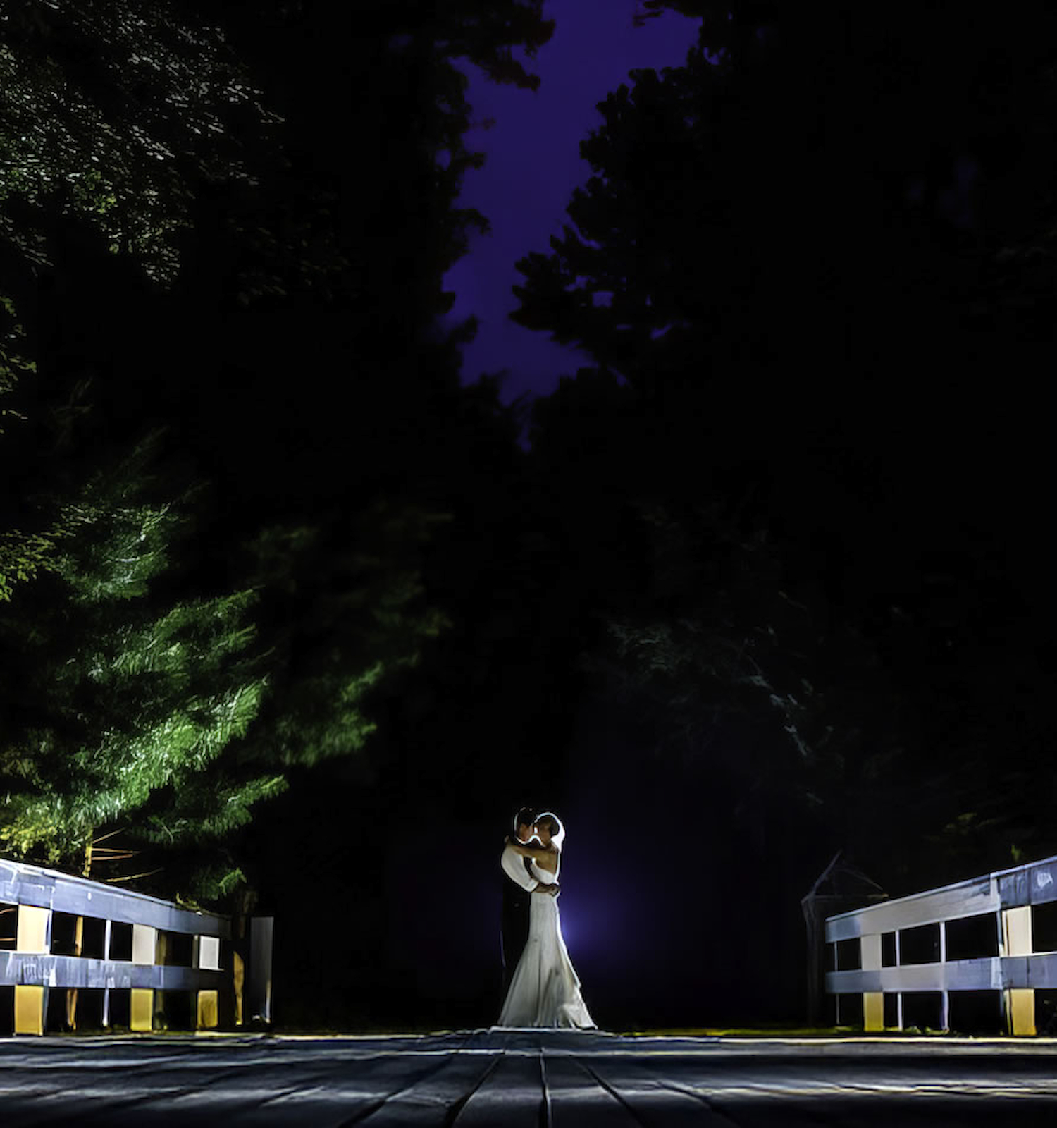 A couple embraces on a wooden bridge lit dramatically against a dark, tree-filled backdrop under a deep purple night sky.