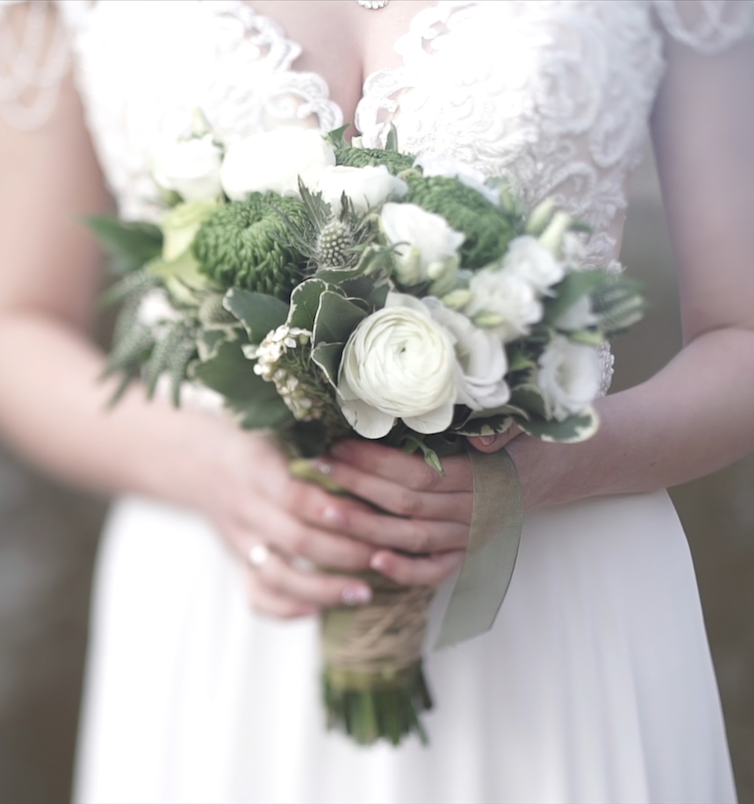 A bride in a wedding dress holds a bouquet of white and green flowers, with a blurred background.