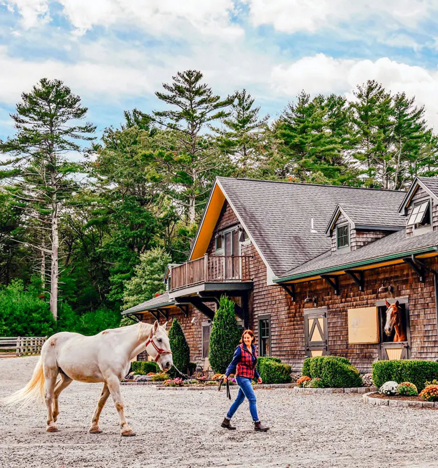 A person is walking a white horse in front of a rustic, wooden building surrounded by trees and greenery, under a partly cloudy sky.