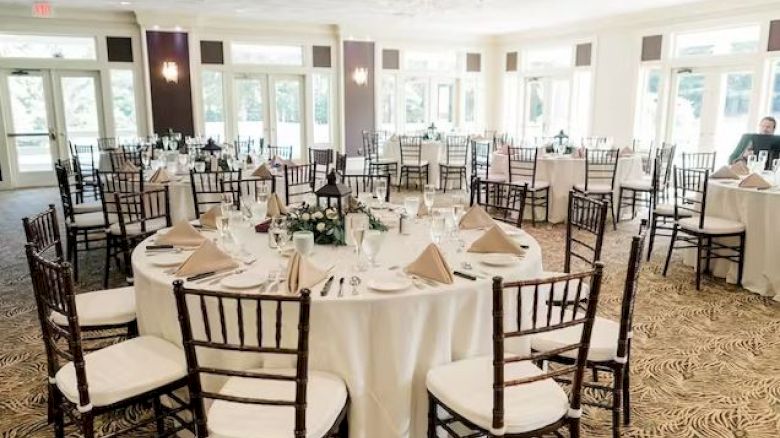 This image shows an elegant banquet hall with round tables covered in white tablecloths, brown chairs, and table settings prepared for an event.