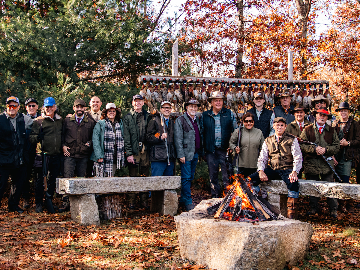 A group of people stand around an outdoor fire pit in a forested area during fall, with benches and a wooden structure in the background.