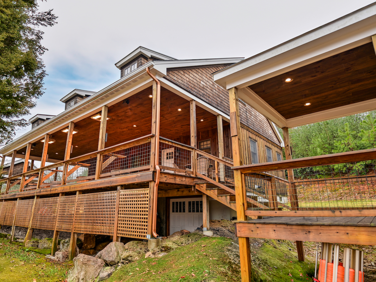 A large wooden house with a spacious porch, multiple columns, and a staircase. Surrounded by greenery and trees, it appears serene.